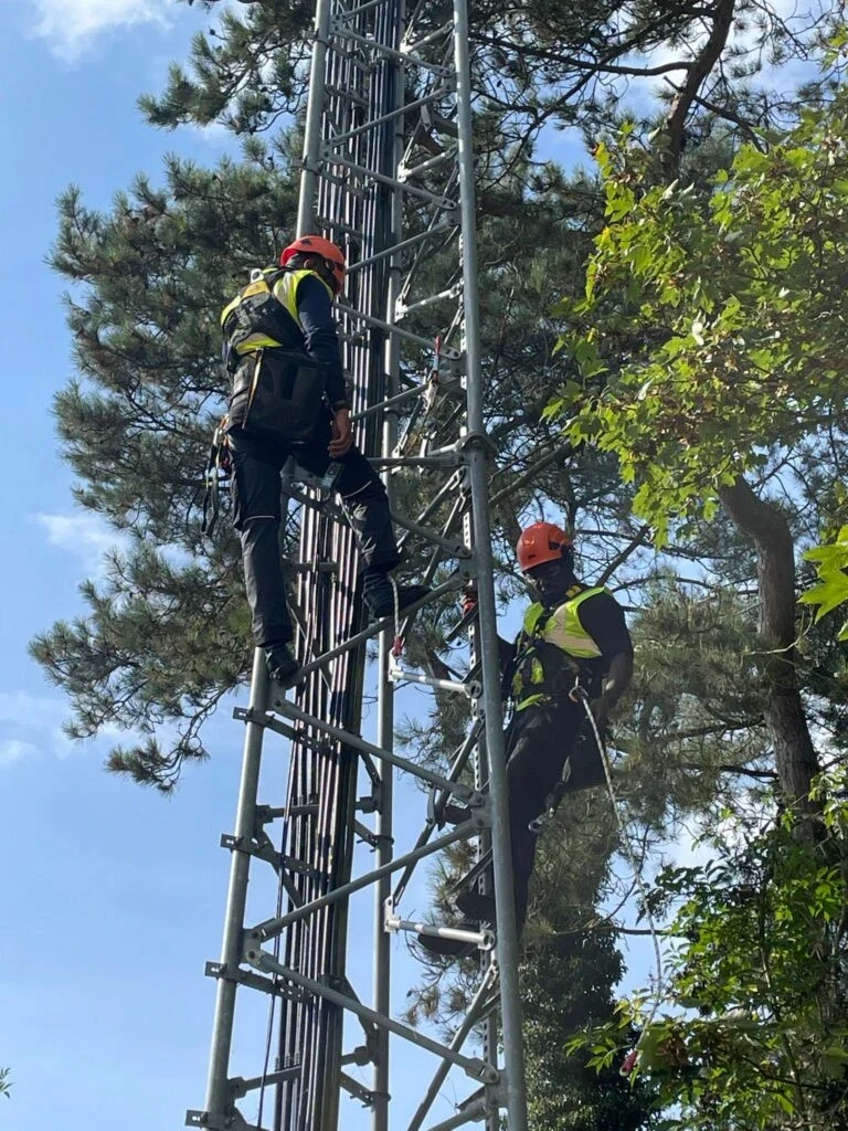 OG Meta riggers in helmets, harnesses and safety vests climbing a lattice tower